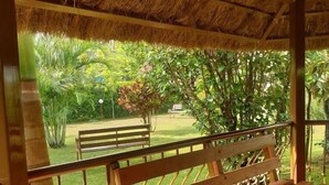 Um banco de madeira sob uma cobertura de palha, com vista para um jardim bem cuidado e outro banco ao longe.