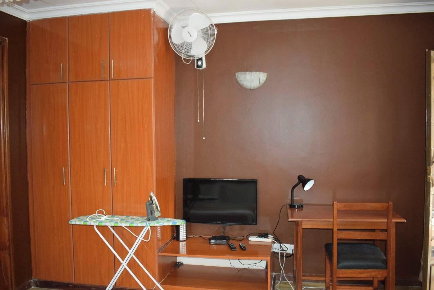 Un espace de vie compact comprenant une armoire en bois, un petit bureau avec une télévision, un ventilateur et une lampe.