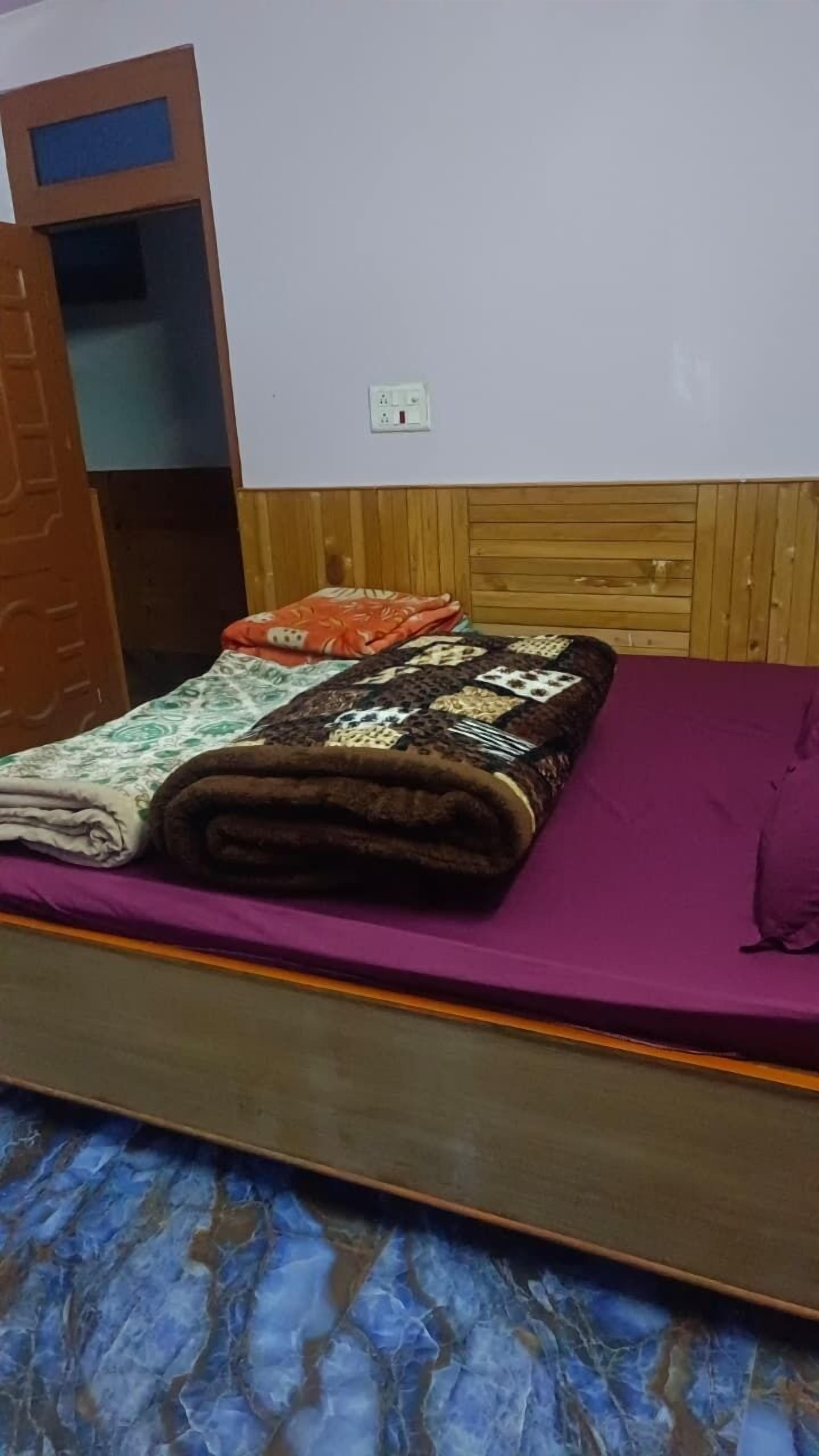 A bed with a purple mattress, a quilt, and a blanket. There is a wooden headboard and a wall socket.