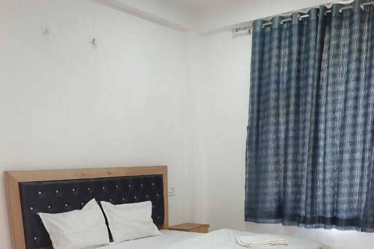 Un dormitorio con estructura de cama de madera, ropa de cama blanca, una mesita de noche pequeña y una ventana con cortinas azules.