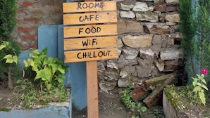 Placa de madeira com informações sobre comodidades, incluindo quartos, café, comida, WiFi e área de relaxamento.