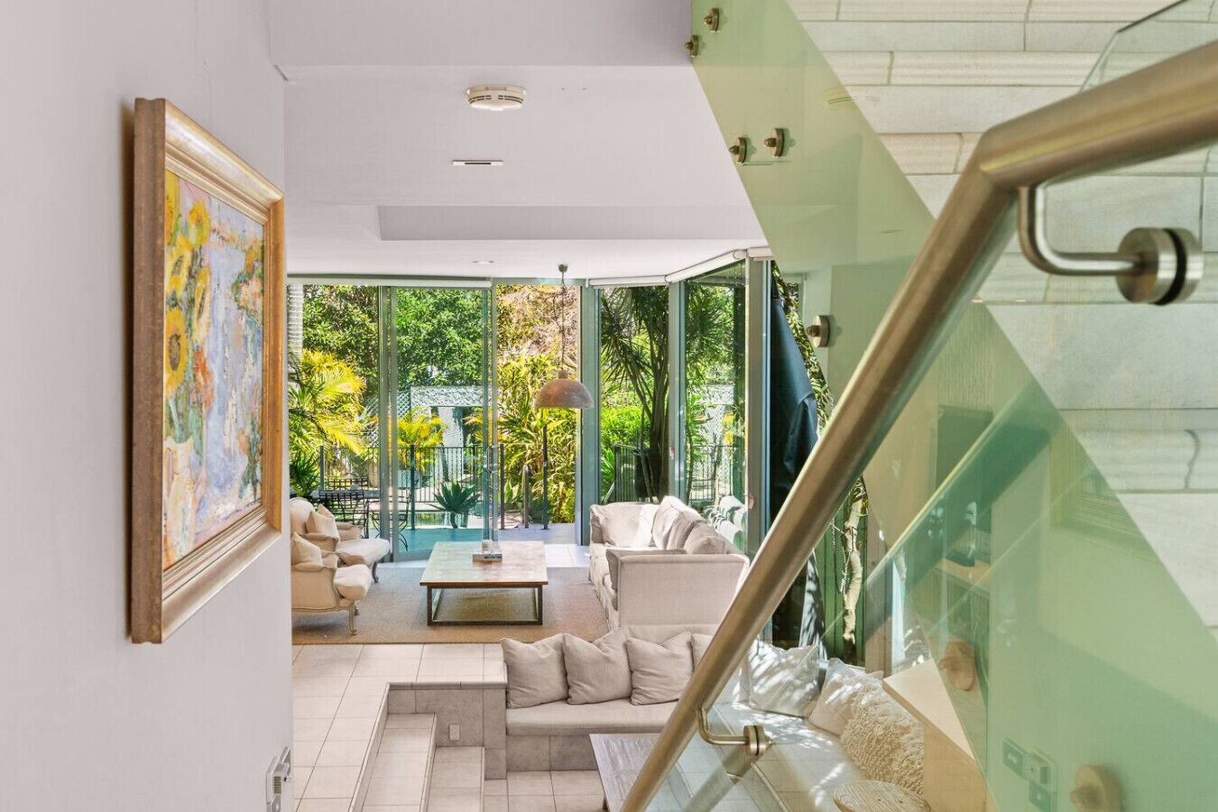 A modern living room with a glass staircase, a painting on the wall, and a view of the outdoors.