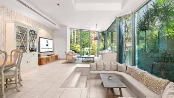 A modern living room with a large sectional sofa, a coffee table, and a TV mounted on the wall. There is a glass door leading to a garden with plants visible outside.