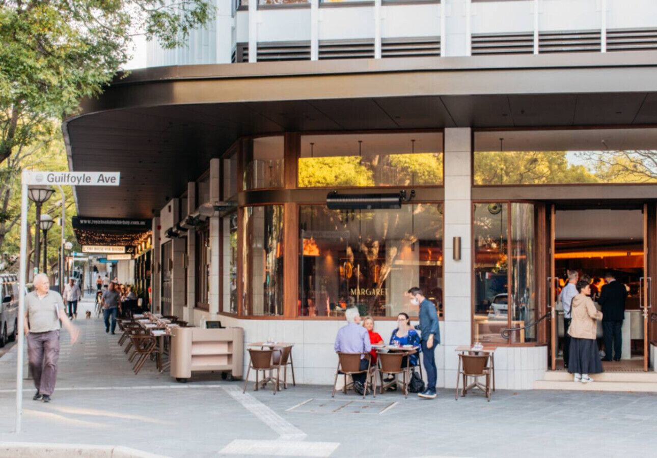 An outdoor cafe area with tables and chairs, a covered seating area, and a building entrance.