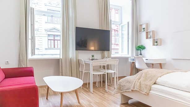 Une chambre d’hôtel moderne avec un lit, un canapé rouge, une table blanche, une télévision et de grandes fenêtres avec des rideaux.