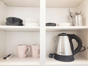 Une cuisine avec des étagères blanches, une bouilloire, deux tasses roses, une cafetière noire et blanche et un récipient en métal contenant des ustensiles.
