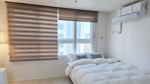 A bedroom with a bed, window blinds, a wall-mounted air conditioner, and a visible cityscape view.