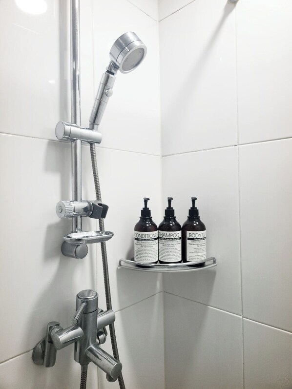A shower with a handheld showerhead and a wall-mounted faucet. There are three bottles of shampoo on a shelf.