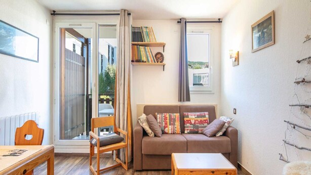 A cozy living room with a brown sofa, a wooden coffee table, and a chair. There's a bookshelf with books and a window with curtains. - Cosy Mountain Retreat for 4 Guests (Villard-de-Lans)