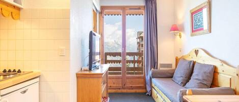 A compact room with a kitchenette, a single bed with a wooden headboard, a small table, and a view of the outdoors through a window with curtains.