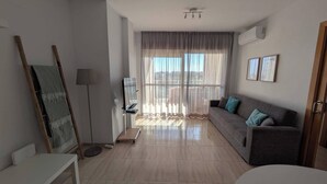 Een moderne woonkamer met een grijze bank, een staande lamp, een flatscreen-televisie en een groot raam met dunne gordijnen.