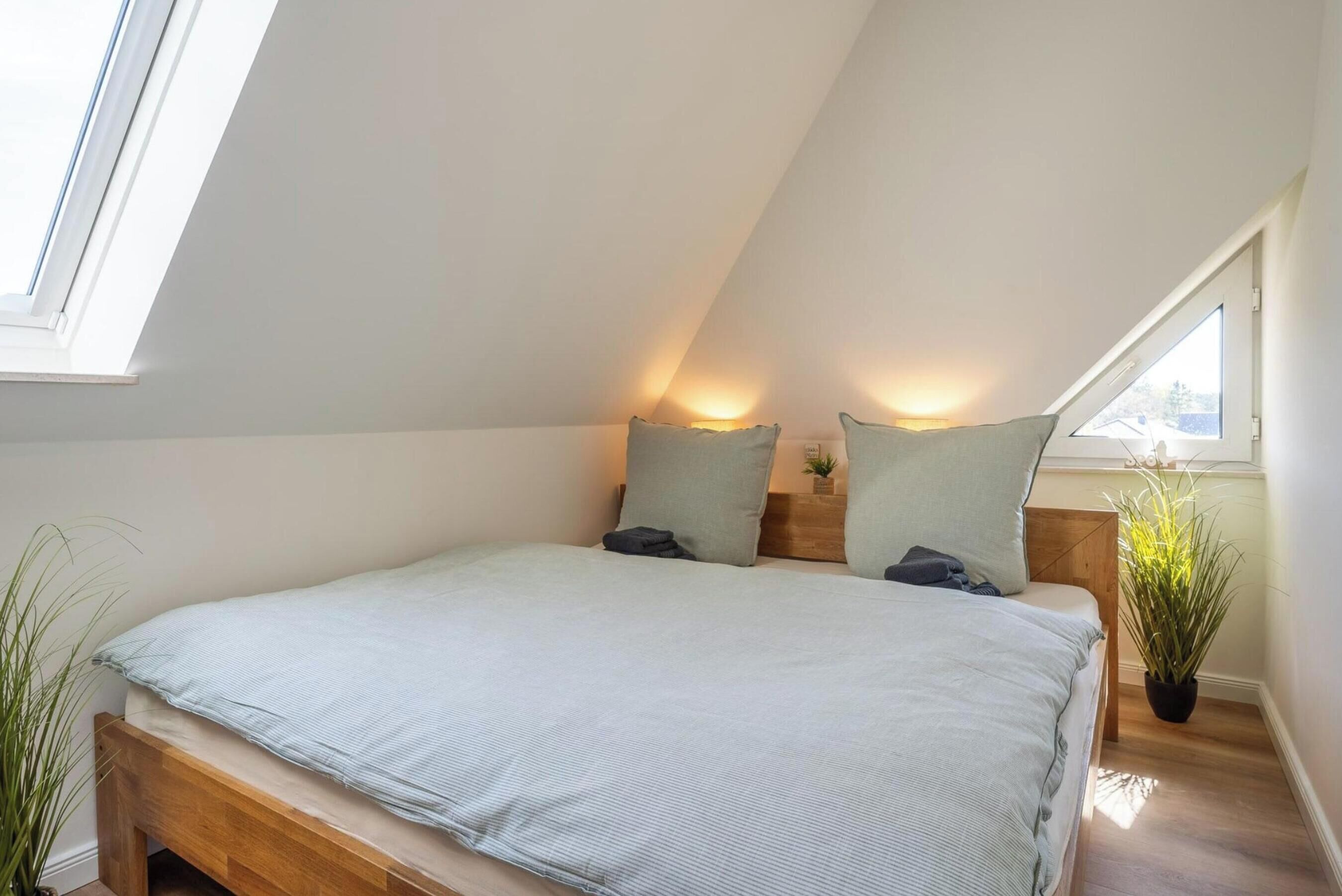 A bedroom with a sloped ceiling, a bed with a light-colored mattress, two pillows, a bedside table with a lamp, and a potted plant.