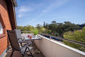 A balcony with a table, chairs, a potted plant, and a railing overlooking a parking area and trees.