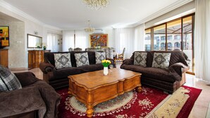 A spacious living room with a large wooden coffee table, two brown sofas with patterned cushions, a chandelier, and a large window with a view of buildings.