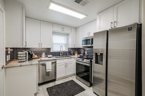 A modern kitchen with white cabinets, stainless steel appliances, a microwave, a refrigerator, and a window with blinds. - Enjoy two separate, fully furnished homes on one property — perfect for large groups. The front one-story home offers 3 bedrooms and 1 bath, while the back two-story home features 3 bedrooms, 2 baths, and an upstairs family room with 2 beds (Houston)