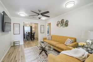 A living room with a sofa, coffee table, and a ceiling fan. There is a dining area in the background. - Enjoy two separate, fully furnished homes on one property — perfect for large groups. The front one-story home offers 3 bedrooms and 1 bath, while the back two-story home features 3 bedrooms, 2 baths, and an upstairs family room with 2 beds (Houston)