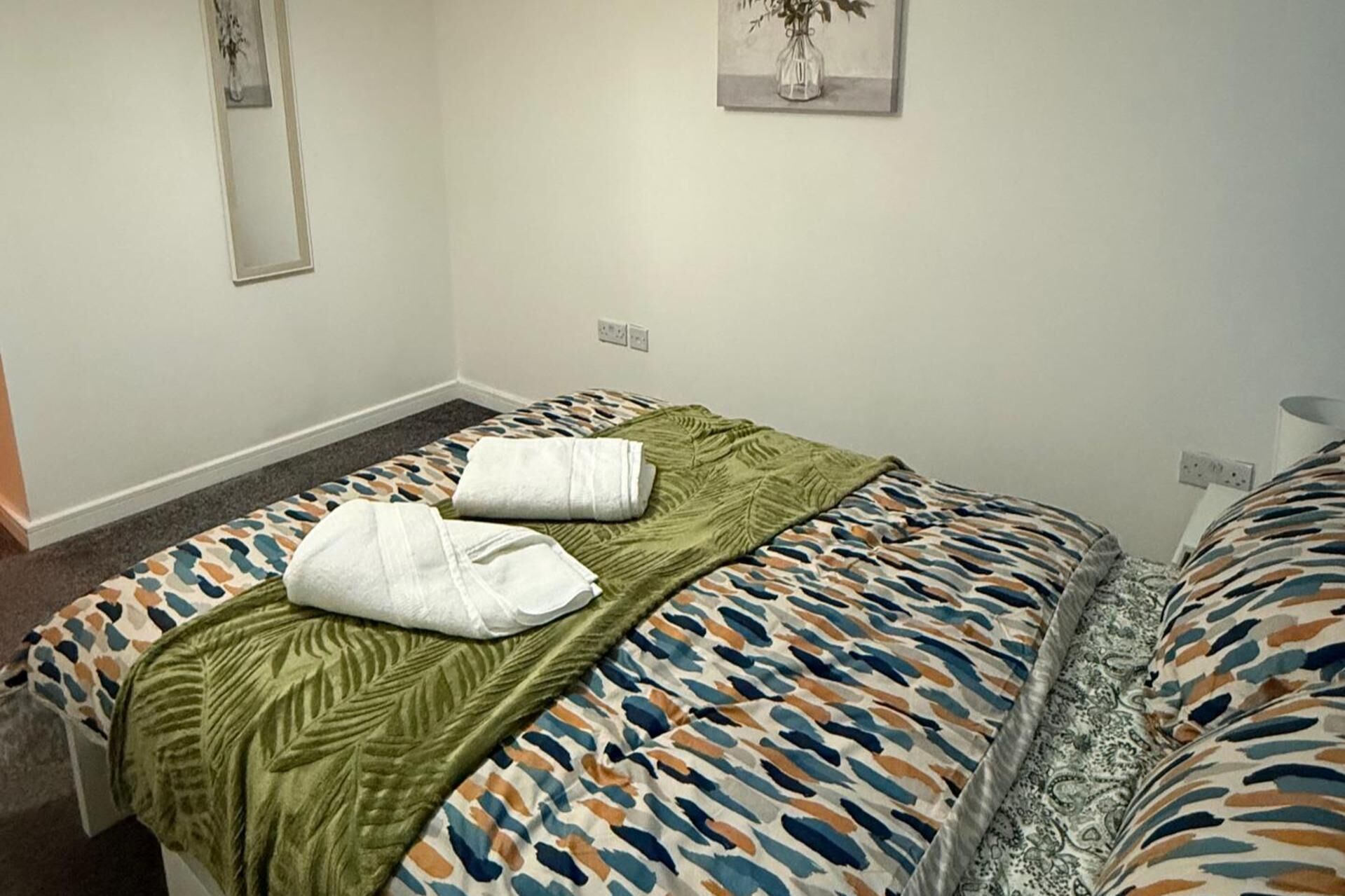 A single bed with a patterned bedspread, a green throw blanket, and two white towels.