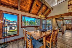 A spacious dining area with a wooden table and chairs, large windows offering mountain views, and a staircase leading to an upper level. - Lakeside Haven by Heavenly Sand (South Lake Tahoe)