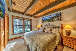 Ein Schlafzimmer mit großem Fenster, einem Bett mit blattgemustertem Überwurf, einem Waldgemälde und einer Holzbalkendecke.