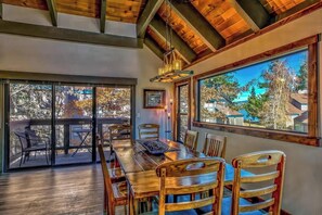 A dining area with a wooden table and chairs, large windows with a view of trees and houses, and a ceiling with exposed beams.