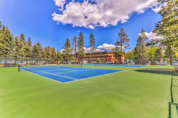 Cancha de tenis al aire libre con una red, rodeada de árboles y un edificio al fondo.