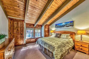 A bedroom with a wooden ceiling, a bed with patterned bedding, a nightstand with a lamp, and a window with a view of trees. - Lakeside Haven by Heavenly Sand (South Lake Tahoe)