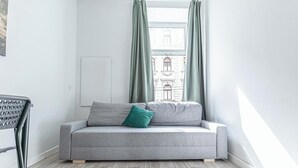 A modern living room with a grey sofa, a green pillow, and a view of a building through sheer curtains.