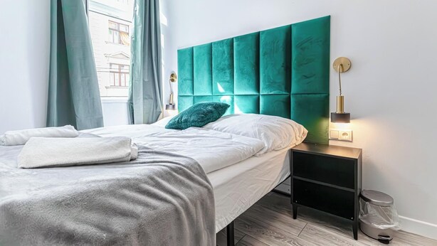 A neatly made bed with a white comforter and a grey blanket, a teal headboard, a bedside lamp, and a small black nightstand with a trash bin.