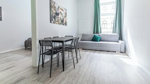 A modern living room with a grey sofa, a dining table with chairs, a painting on the wall, and a window with curtains.