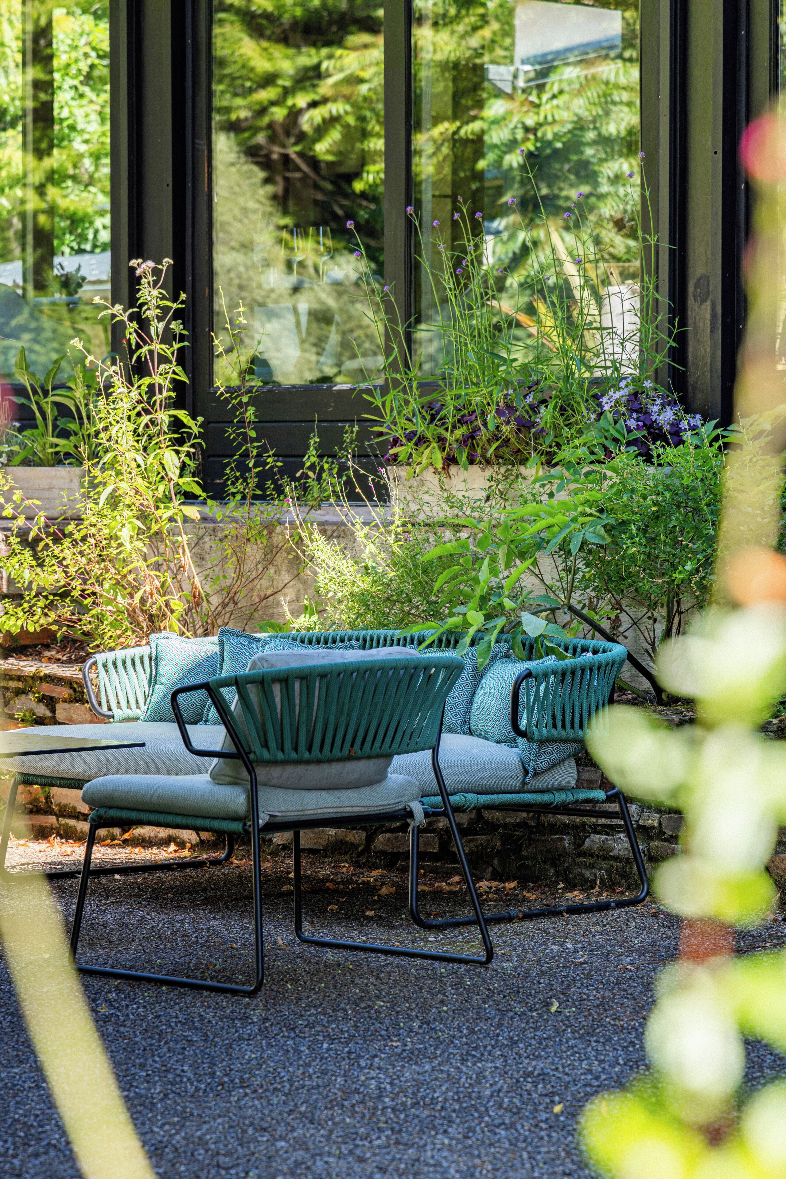 Un espace extérieur verdoyant avec des sièges, un banc et des coussins, entouré de plantes et d’un mur en pierre.