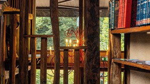 A rustic wooden interior with a wooden staircase leading to a balcony, a bookshelf filled with books, and a view of greenery outside. - Le Moulin de Cambelong (Conques-en-Rouergue)