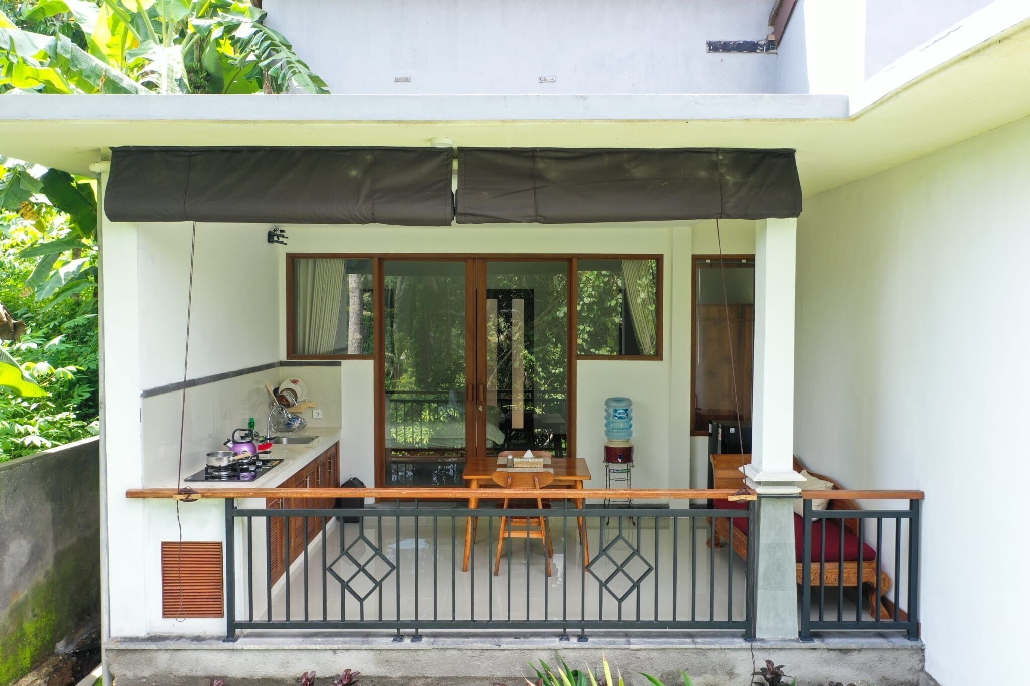 A small balcony with a wooden table and chairs, a water dispenser, and a view of the surrounding greenery.