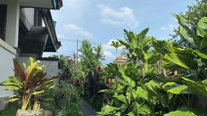 A narrow pathway flanked by lush greenery and potted plants, leading to a building with a visible staircase and a clear sky. - Vishnu Villa (Ubud)