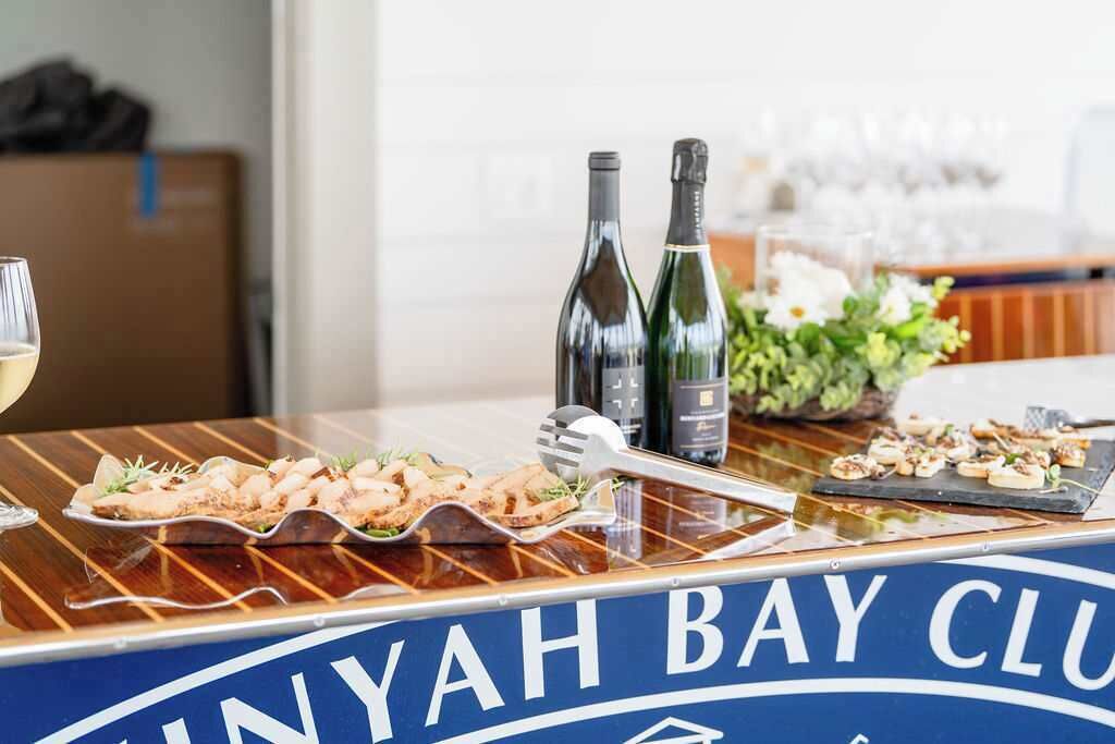 Een bar met een bord waarop 'YAH BAY CLUB' staat, een fles wijn en een wijnglas op de toonbank.