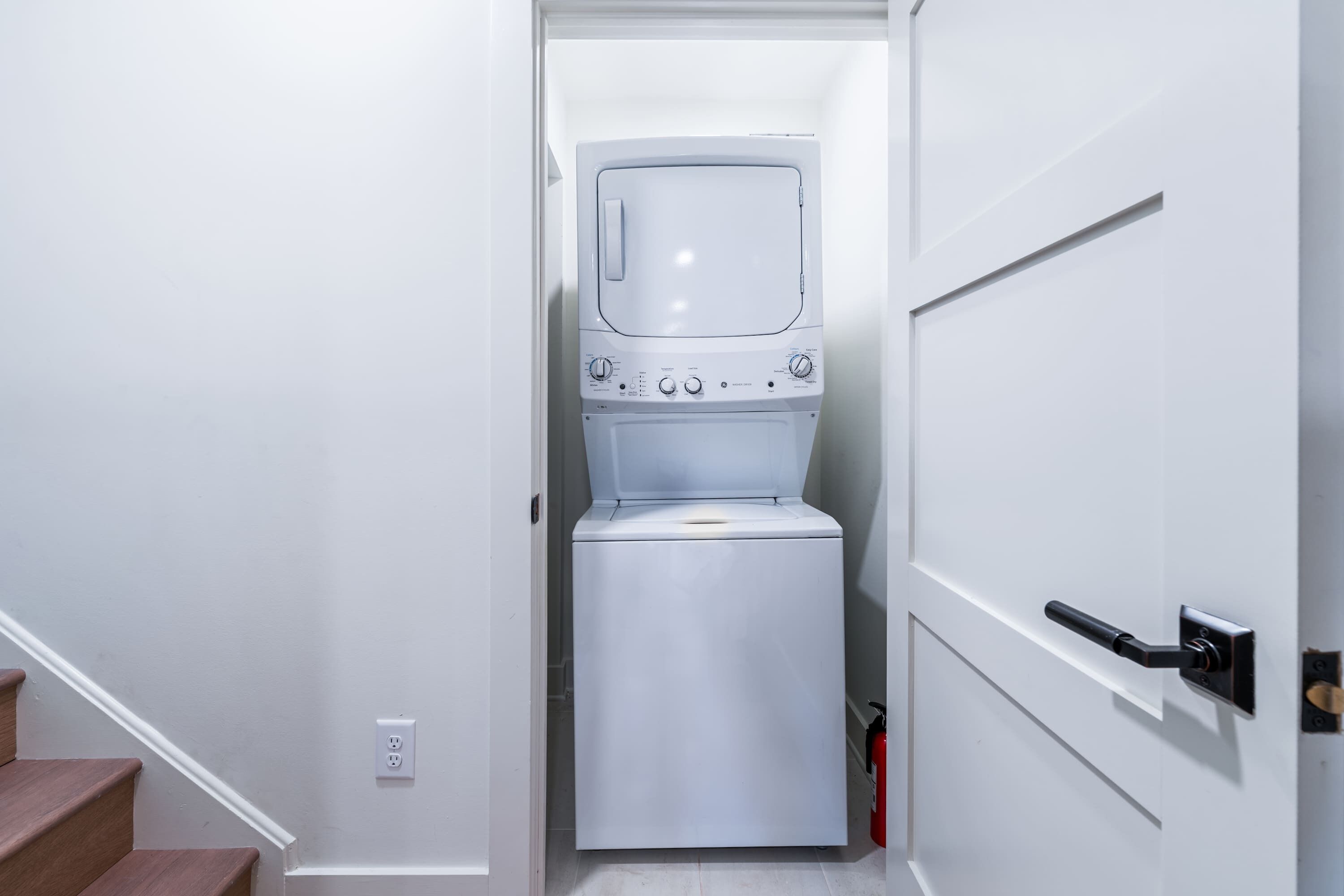 Un lavadero pequeño con lavadora y secadora, una puerta con manija y una escalera que lleva a otra habitación.