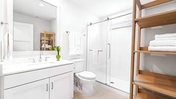 Un baño moderno con tocador blanco, un espejo grande, inodoro, cabina de ducha con mampara de vidrio y estante de madera con toallas.