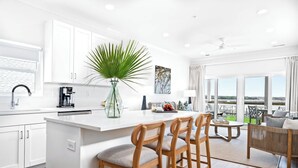 Una cocina moderna con muebles blancos, una isla central con taburetes de bar, una planta grande en maceta y un comedor con vistas a la ciudad.