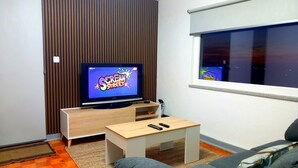 Una sala de estar moderna con un televisor de pantalla plana mostrando 'Scream Street', un mueble de televisión de madera, una pequeña mesa de madera con dos mandos a distancia, un sofá y una ventana con vistas al cielo nocturno.