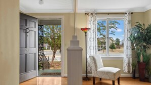 Une pièce avec un plancher en bois, une chaise blanche, une haute colonne blanche, une fenêtre avec des rideaux blancs et une vue sur l’extérieur.