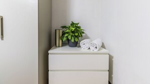A white bedside table with a potted plant, two rolled towels, and a book.