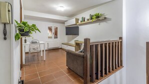 A modern living room with a sofa, a dining table with chairs, a TV mounted on the wall, and a staircase with wooden railings.