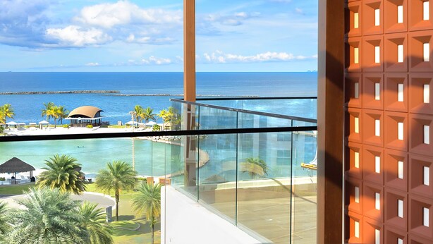 A resort with a glass balustrade overlooking a pool and ocean, palm trees, and a beach area with umbrellas.