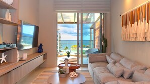 A modern living room with a large TV, a sofa, a coffee table with decorative items, and a view of the ocean through large glass doors.