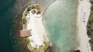 An aerial view of a beach resort with a sandy beach, a pool area with lounge chairs, and a small building.