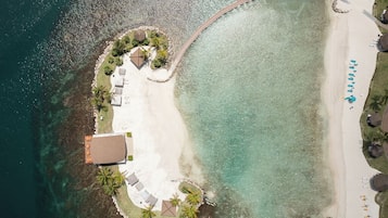 Een luchtbeeld van een strandresort met een zandstrand, een zwembadgedeelte met ligbedden en een klein gebouw.