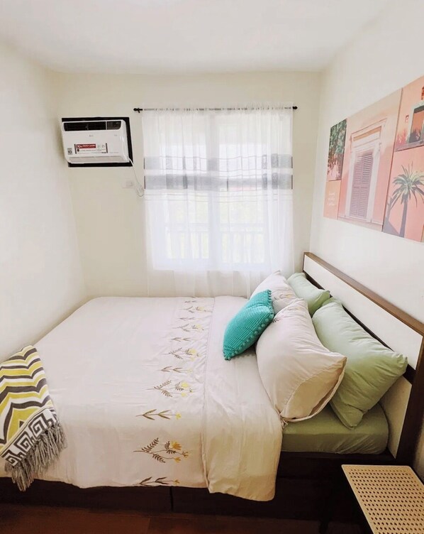A bedroom with a bed, a window with sheer curtains, a wall-mounted air conditioner, and a framed picture.