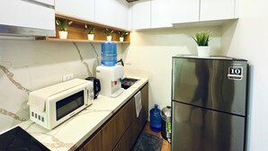 A compact kitchen with white cabinets, a stainless steel refrigerator, a microwave, a water dispenser, and a few potted plants on the countertop. - Cozy 2BR Condo at Seawind Davao • Smart Lock • Netflix 4K & YouTube Premium (Davao City)