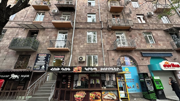 Un edificio de varios pisos con balcones, un restaurante visible con su menú de comida y un supermercado cercano.