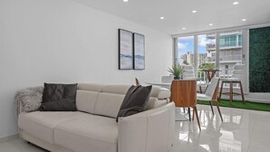 Een moderne woonkamer met een beige bank, een kleine houten tafel en witte stoelen. Er is een groot raam met uitzicht op gebouwen en een groene wand.