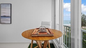 Een ronde houten tafel met een dienblad en peper- en zoutstel, een witte stoel, en uitzicht op het strand door grote ramen.